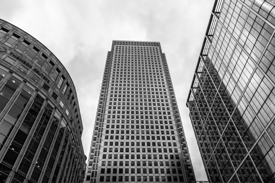 A cityscape scene during the evening featuring a row of modern high-rise office buildings with glass and steel exteriors, illuminated by interior and exterior lighting. The buildings vary in height an