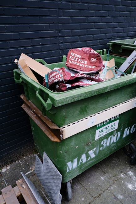 A large green plastic waste bin filled with red and black polypropylene bags of building or garden materials, including a bag labeled 'FK200 FLEXI PLUS FLEXIBLE LEADER CZTE S1,' positioned beside a bl