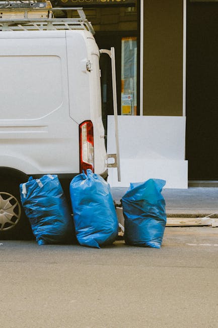A small dark red flatbed truck parked on a city street, loaded with large black, white, and grey bags of waste material. The bags appear to be full and are made of plastic or woven fabric, with some s